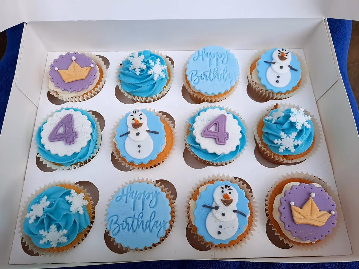 A dozen Frozen-themed birthday cupcakes presented in a white box. The cupcakes feature light blue frosting and fondant decorations including Olaf, white snowflakes, the number four, 'Happy Birthday' text, and a golden crown on purple.