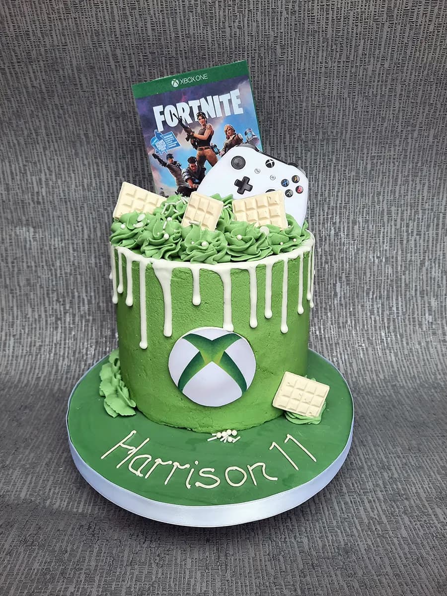 Round green cake with white drip icing, topped with green piped frosting, white sprinkles, white chocolate bars, an Xbox controller, and a Fortnite game cover. The side features an Xbox logo, and the green cake board is decorated with 'Harrison 11' in white icing.