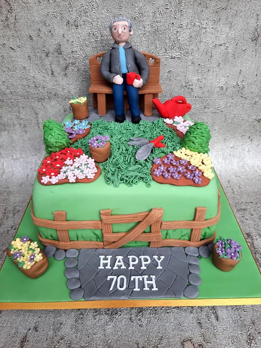A multi-tier green birthday cake decorated with a detailed garden theme, featuring a figure of an older man with glasses sitting on a wooden bench, surrounded by various potted flowers, gardening tools, and a wooden fence, with a paved path at the base reading 'HAPPY 70TH'.