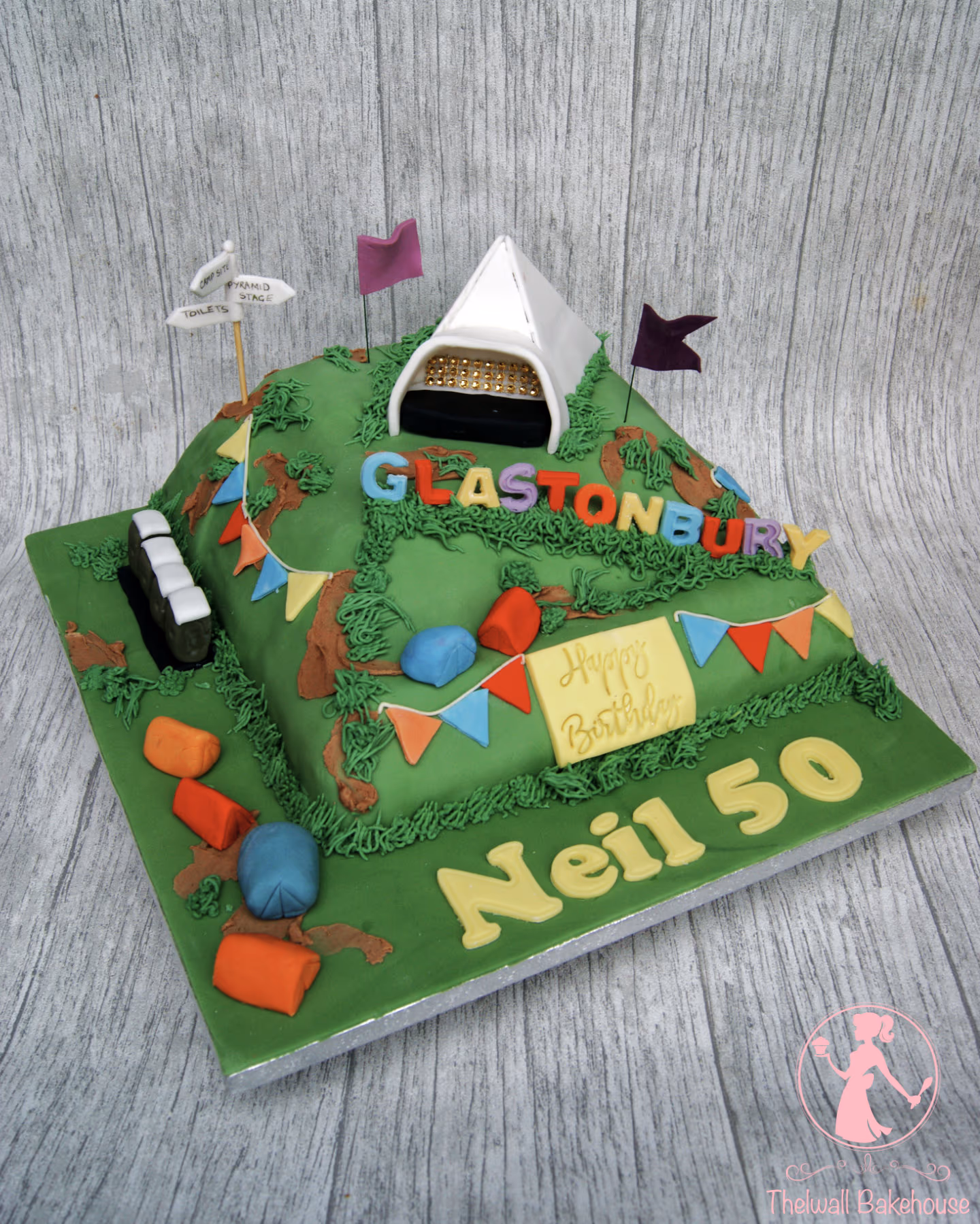 A multi-level green cake decorated to resemble the Glastonbury Festival, featuring a white Pyramid Stage tent, colorful bunting, smaller tents, flags, and text that reads 'Glastonbury', 'Happy Birthday', and 'Neil 50'.