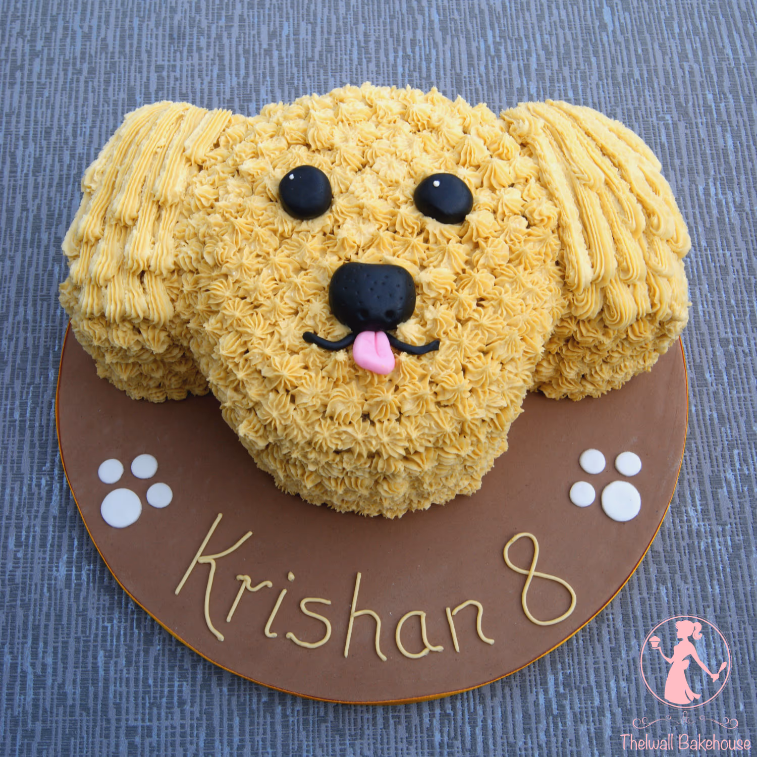 A golden retriever dog head shaped birthday cake, decorated with yellow piped buttercream frosting, black eyes and nose, and a pink tongue, on a brown cake board personalized with 'Krishan 8' and white paw prints.