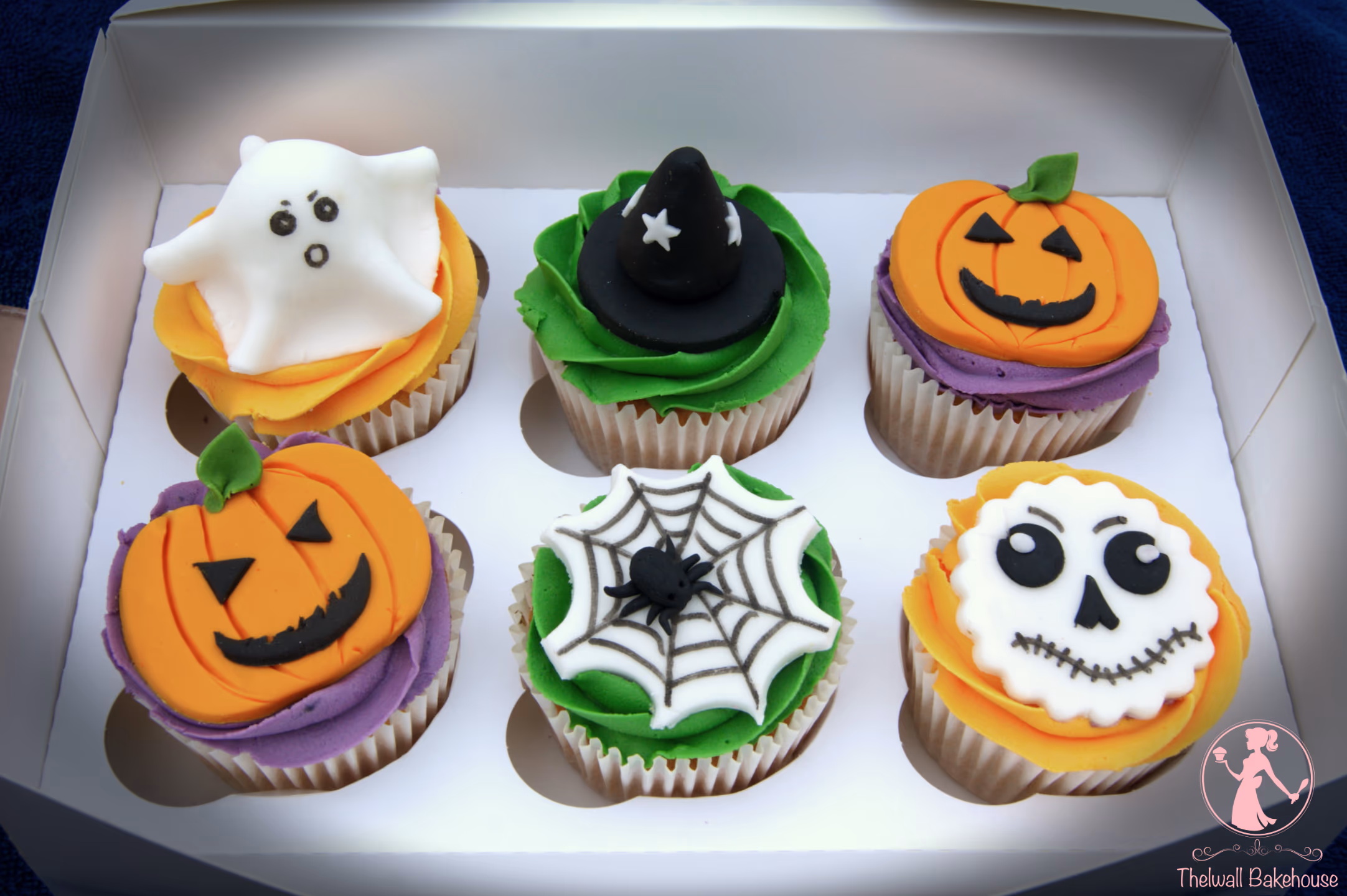 A white box containing six Halloween-themed cupcakes. The cupcakes feature decorations of a white ghost, a black witch hat, two orange jack-o'-lanterns, a white spiderweb with a black spider, and a white skull with black features, all on various colored frostings.