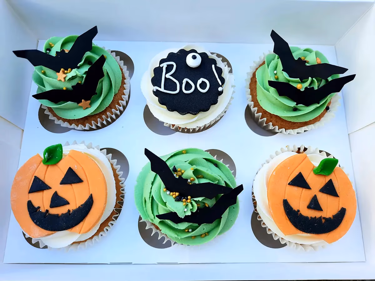 A box of six Halloween-themed cupcakes, featuring designs with green frosting and black bats, white frosting with orange jack-o'-lanterns, and a black 'Boo!' disc with a white eyeball.