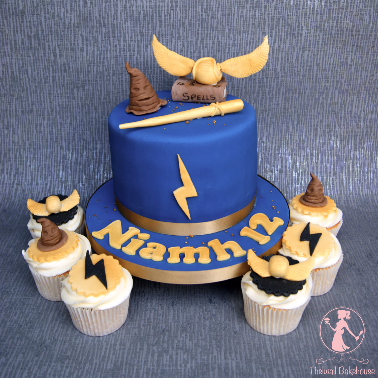 A Harry Potter themed birthday cake and matching cupcakes. The main round cake is dark blue with a gold base, decorated with 'Niamh 12' in gold letters and a gold lightning bolt. On top are a brown sorting hat, a golden wand, and a golden snitch resting on a brown 'SPELLS' book. Surrounding the cake are several cupcakes, each topped with either a brown sorting hat, a golden snitch, or a black lightning bolt.