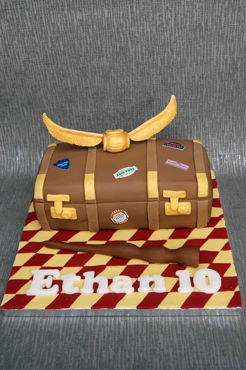 A Harry Potter themed cake shaped like a brown suitcase, decorated with a golden snitch on top, a brown wand, and the name 'Ethan 10' in white letters on a red and yellow checkered base.