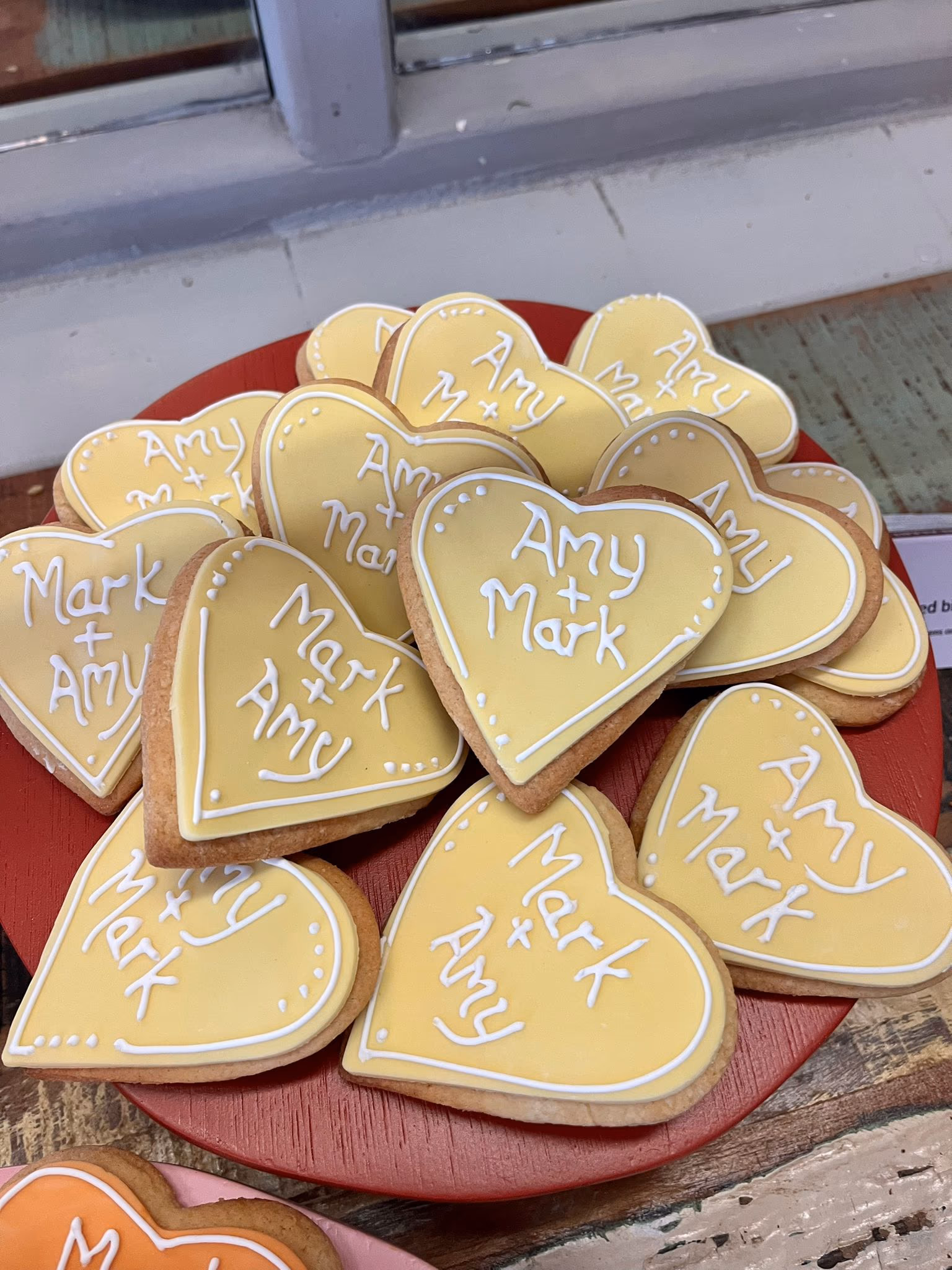 A pile of heart-shaped biscuits with yellow icing, each decorated with 'Mark + Amy' or 'Amy + Mark' in white frosting, displayed on a red platter.