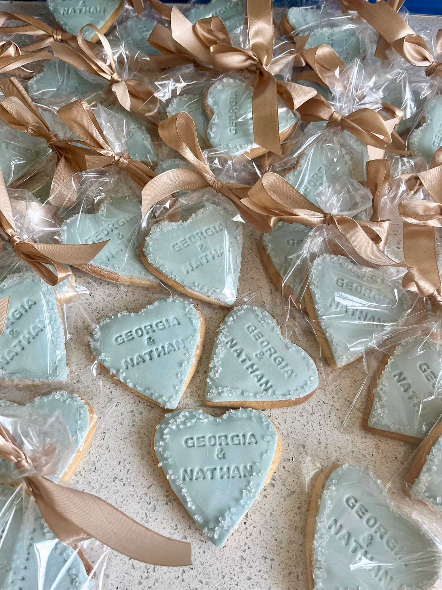 Close-up view of numerous heart-shaped cookies with light blue icing, personalized with 'Georgia & Nathan' and decorated with white sprinkles, most individually wrapped in clear plastic bags tied with gold ribbons.