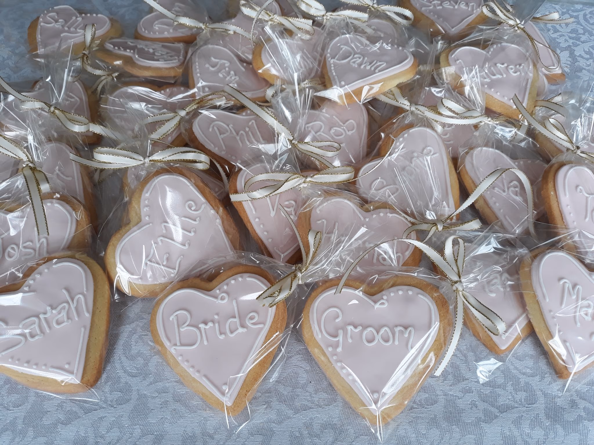 A collection of individually wrapped heart-shaped biscuits with pink icing and white personalized names, tied with white and gold ribbons, displayed as wedding favors.