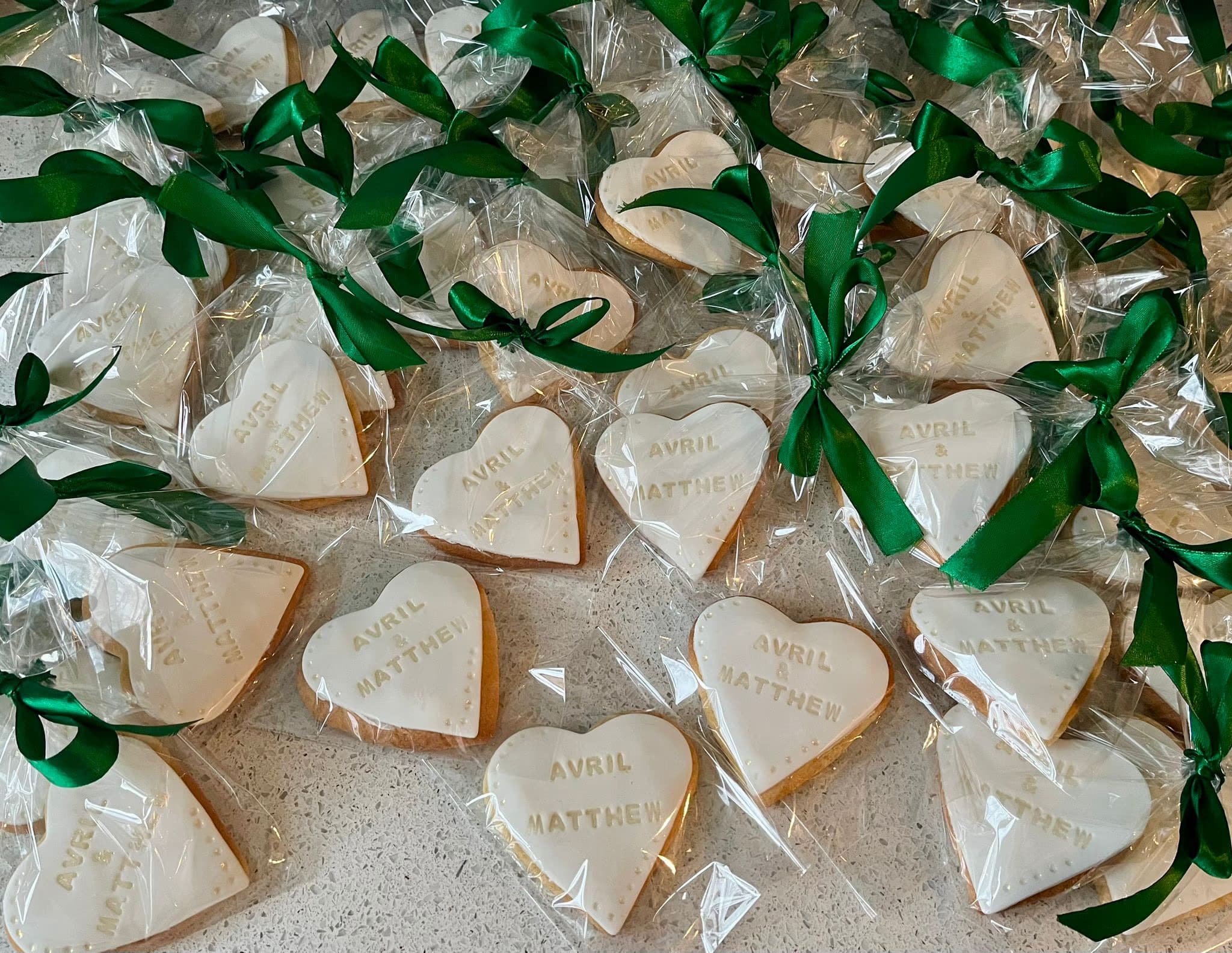 A flat lay of many heart-shaped white-iced biscuits, each individually wrapped in clear plastic and tied with a green satin ribbon. The biscuits are personalised with the names 'Avril & Matthew' in gold lettering, and some have small decorative dots around the edges.