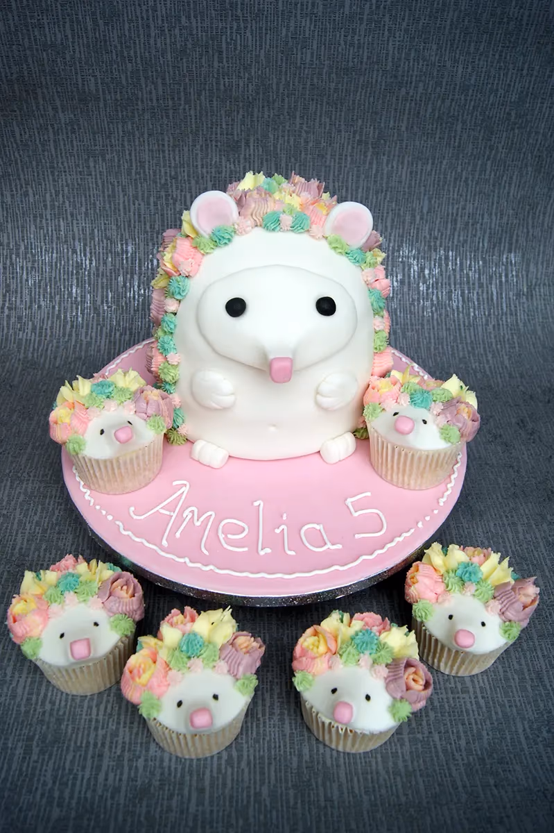A white hedgehog-shaped birthday cake with pastel pink, yellow, green, and purple frosting spikes, pink ears, and a pink nose, sitting on a pink cake board that says 'Amelia 5'. The cake is surrounded by six matching hedgehog cupcakes with similar pastel frosting.