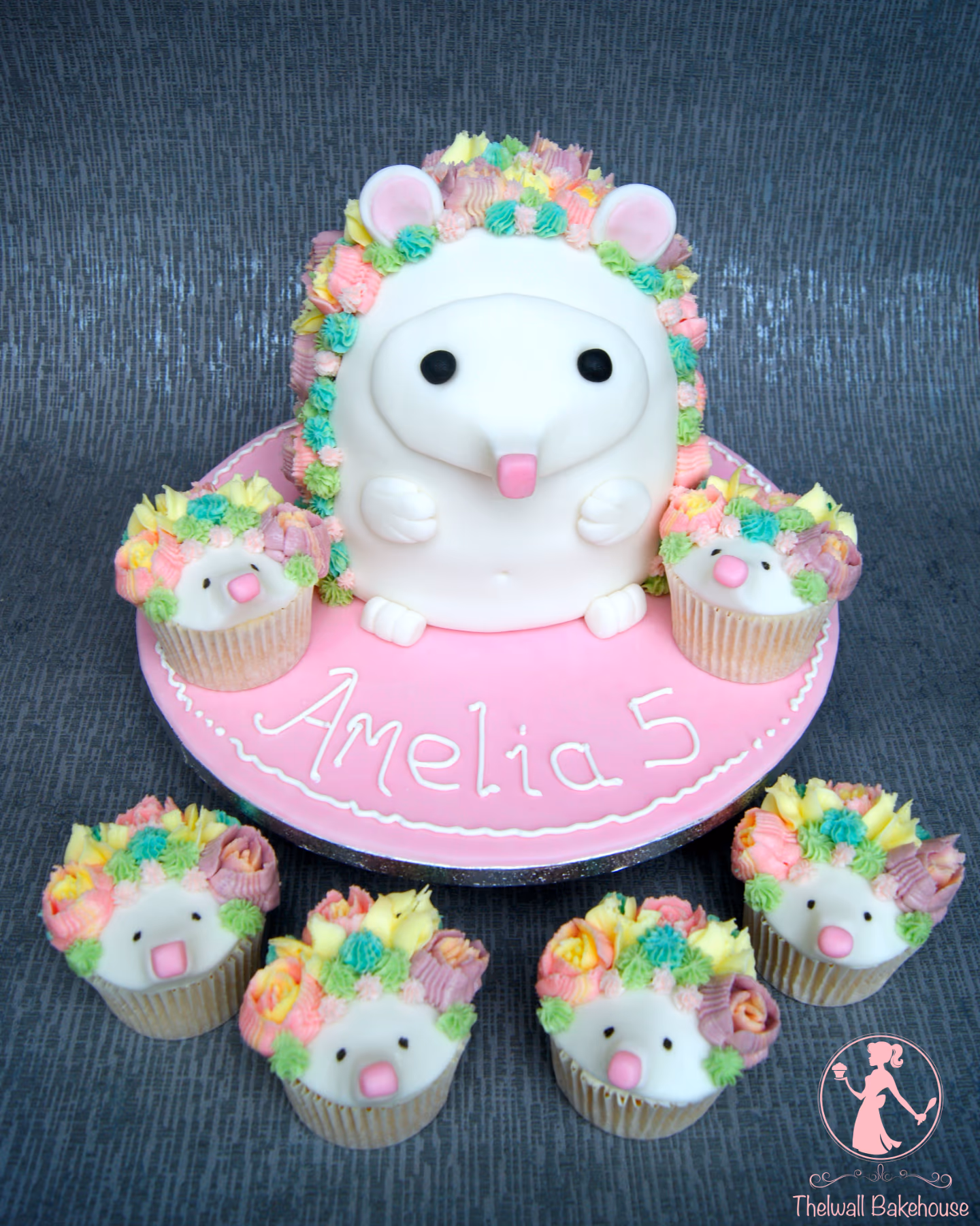 A large white fondant hedgehog birthday cake with pastel pink, yellow, green, and purple buttercream spikes, sitting on a pink cake board with "Amelia 5" written in white. Six matching hedgehog cupcakes with pastel buttercream spikes are arranged around the main cake.