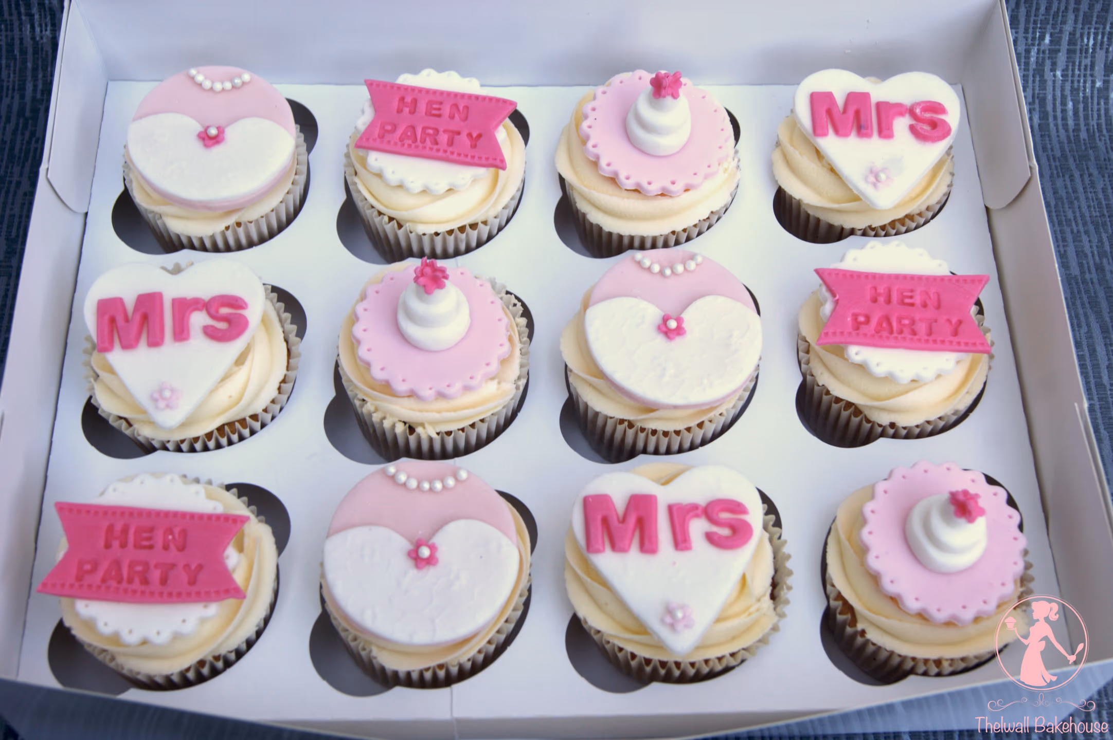 A box of twelve hen party themed cupcakes with pink and white fondant decorations, including 'Mrs' hearts, 'Hen Party' banners, bridal dress designs, and fascinator designs.