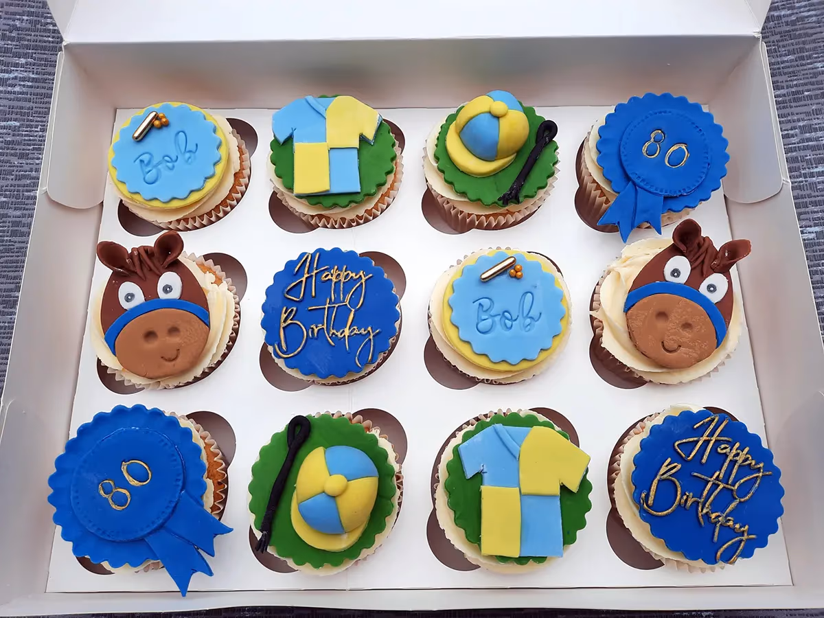 Twelve horse racing themed cupcakes in a white box for an 80th birthday, featuring designs of horse heads, jockey shirts, jockey hats with whips, blue rosettes with '80', and messages like 'Happy Birthday' and 'Bob'.