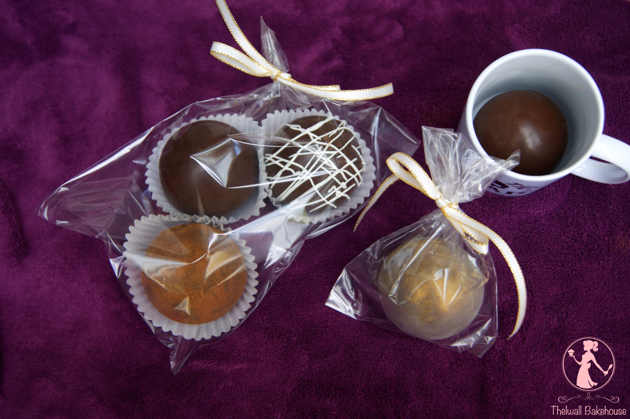 Assorted hot chocolate bombs are displayed on a purple background, with three in one clear bag (two dark chocolate, one with white drizzle, one golden), one golden bomb in another clear bag, and a dark chocolate bomb in a white mug.