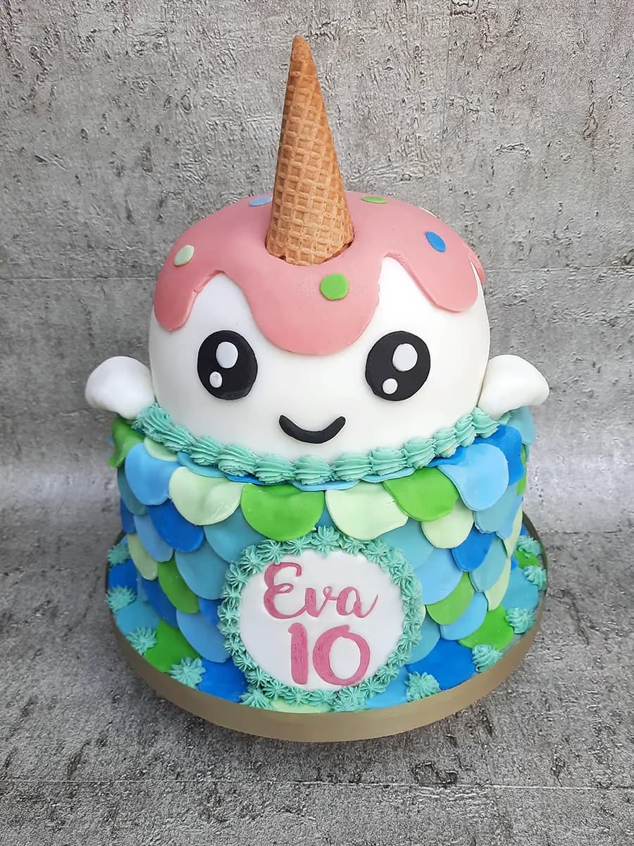 A two-tier birthday cake featuring a cute white character with a pink ice cream drip and an ice cream cone horn on the top tier, and a bottom tier decorated with blue and green mermaid-like scales, with a white plaque displaying 'Eva 10' in pink.
