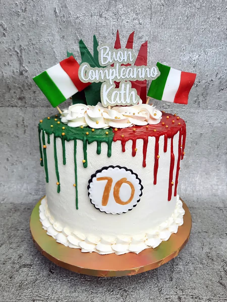 A white birthday cake decorated with Italian flag colors, featuring green and red drip icing on top, two miniature Italian flag toppers, a 'Buon Compleanno Kath' topper, and the number '70' on a white and black scalloped disc on the side.