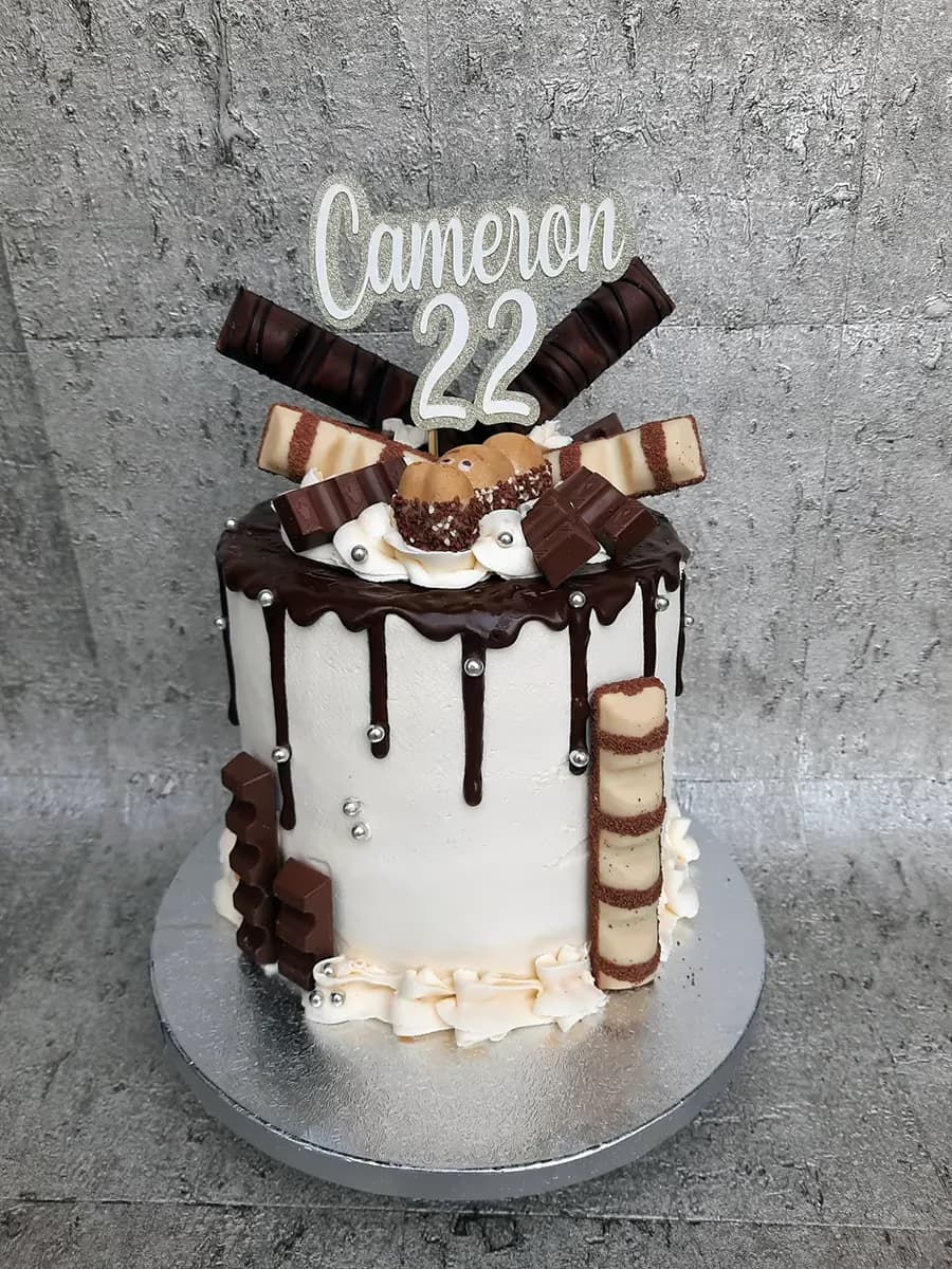 A white birthday drip cake decorated with chocolate ganache, various Kinder chocolate bars, and silver sprinkles, topped with a 'Cameron 22' topper.