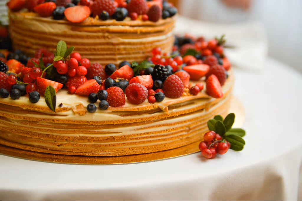 A two-tier layered cake, possibly a honey or crepe cake, generously topped with a vibrant assortment of fresh mixed berries including strawberries, raspberries, blueberries, and redcurrants.