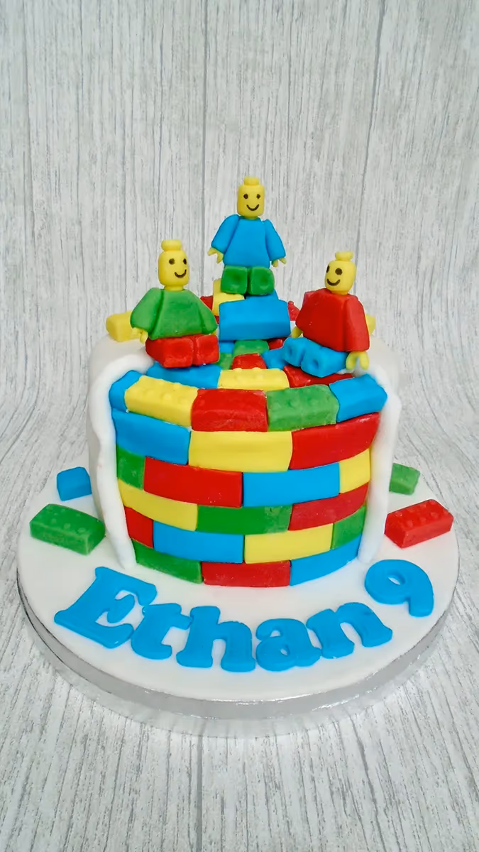 A round single-tier cake decorated with colorful fondant bricks in red, blue, green, and yellow, resembling Lego bricks. Three Lego-style minifigures stand on top, and the name 'Erhan 9' is written in blue fondant on the white cake board.