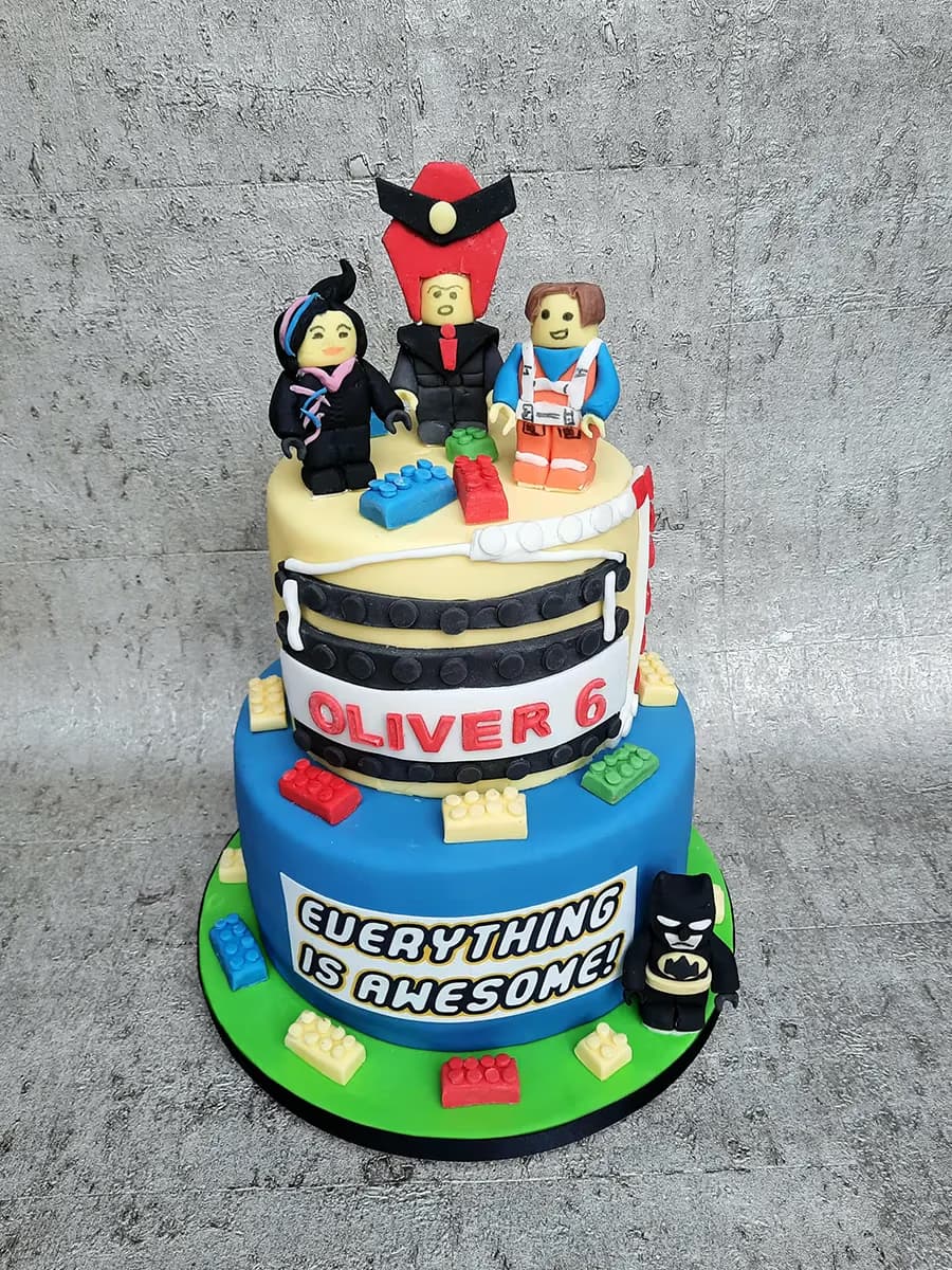 A two-tier Lego Movie themed birthday cake for Oliver's 6th birthday, featuring fondant figures of Emmet, Wyldstyle, Lord Business, and a small Batman, with colorful Lego brick decorations and the text 'OLIVER 6' and 'EVERYTHING IS AWESOME!'.