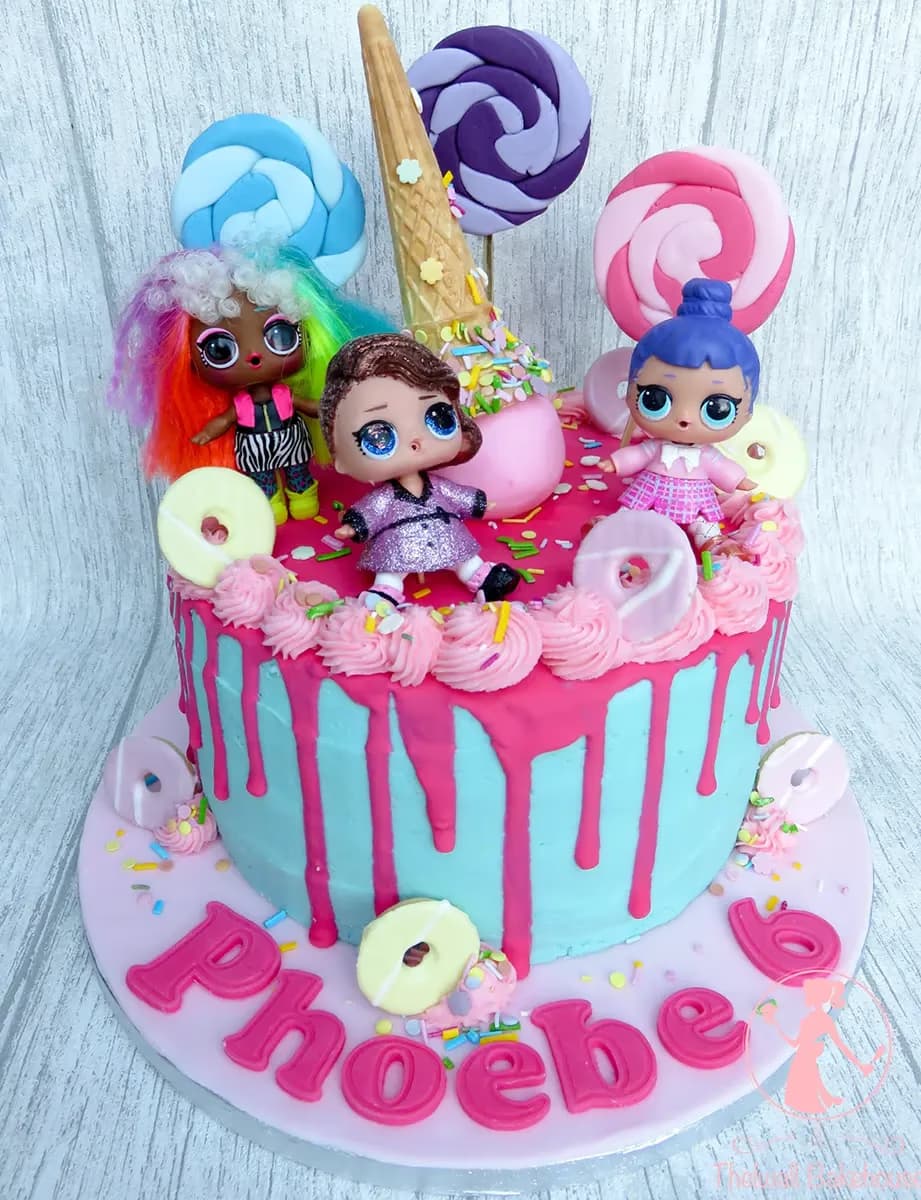 A vibrant birthday cake with light blue frosting and bright pink drip icing, topped with three LOL Surprise dolls, colorful lollipop decorations, an ice cream cone, and sprinkles, with the name Phoebe and the number 6 on the cake board.