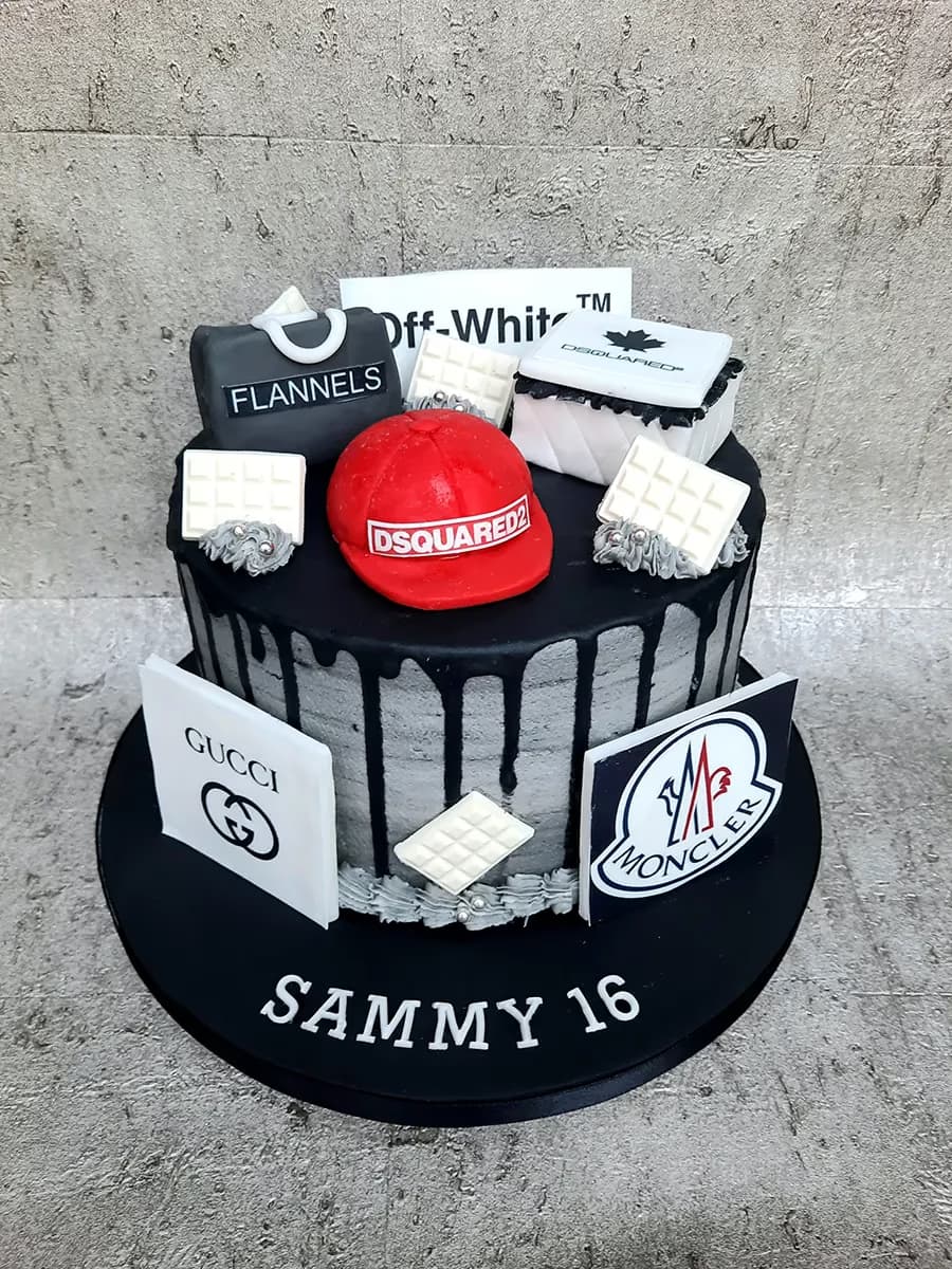 A single-tier birthday cake decorated with luxury fashion brand logos and items, including Off-White, Flannels, Dsquared2, Gucci, and Moncler, featuring black drip icing over grey textured sides, and 'SAMMY 16' written on the black cake board.