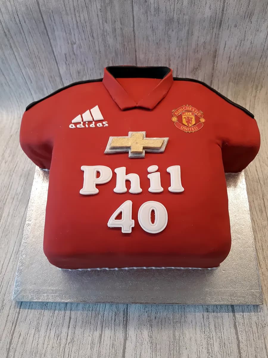 A red football shirt shaped cake, decorated with a Manchester United crest, an Adidas logo, a gold Chevrolet logo, and the name 'Phil' and number '40' in white icing.