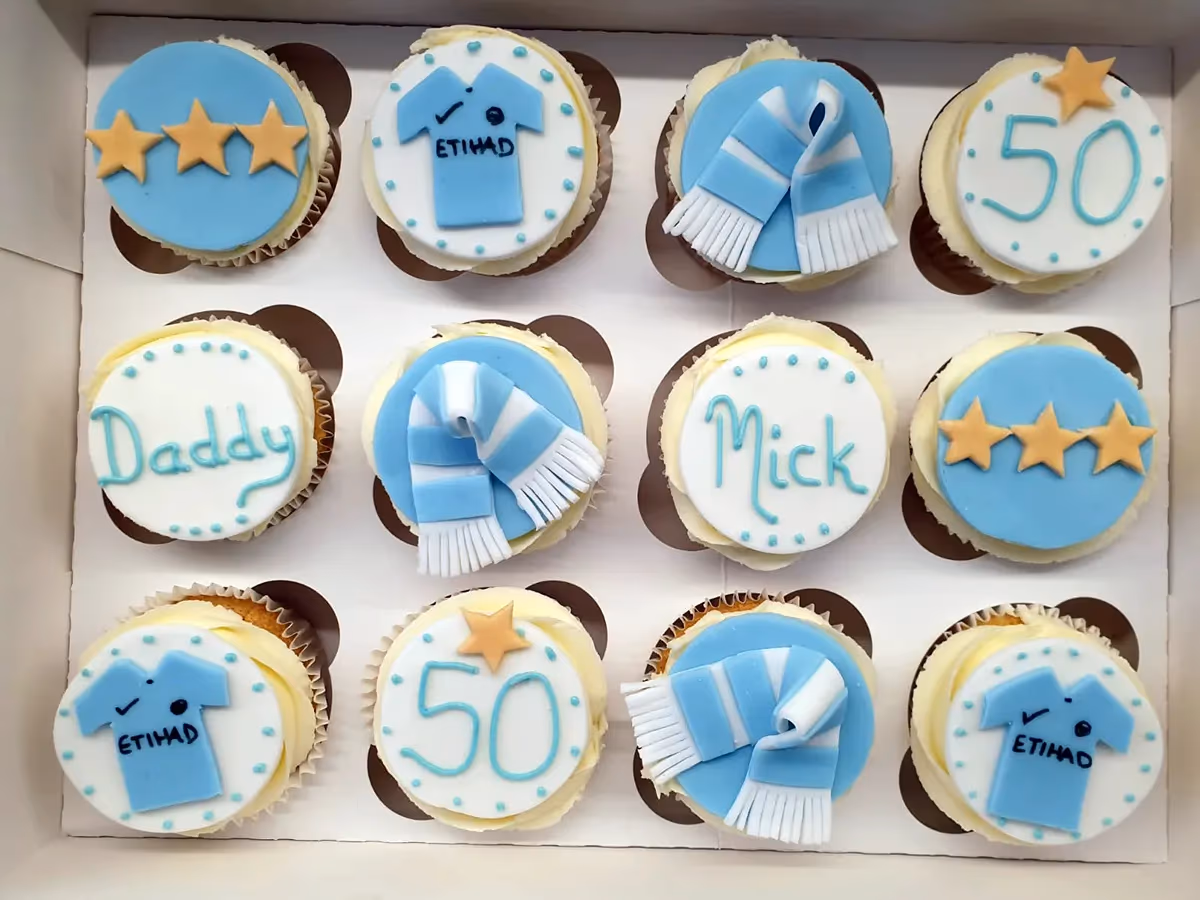 A box of twelve Manchester City themed cupcakes with light blue and white fondant decorations, including football jerseys with 'ETIHAD', striped scarves, the number '50', gold stars, and the names 'Daddy' and 'Mick'.