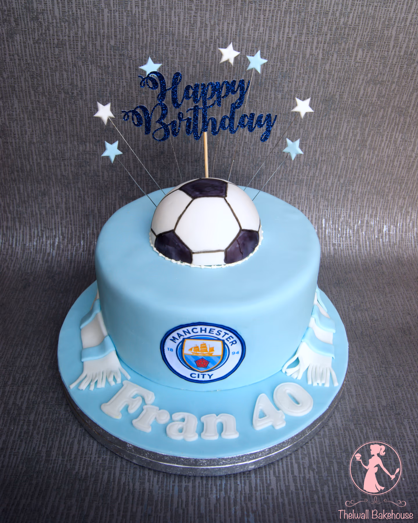 A light blue round birthday cake with a Manchester City theme, featuring a fondant football on top, the Manchester City logo on the side, a light blue and white scarf draped around the base of the cake, and 'Fran 40' written on the cake board, topped with a blue glitter 'Happy Birthday' sign and white and light blue stars.
