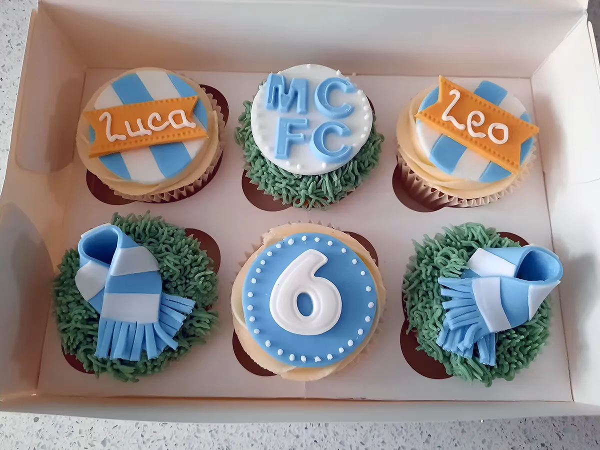 Six Manchester City themed cupcakes in a white box, featuring names 'Luca' and 'Leo', 'MCFC' logo, a number '6', and blue and white striped scarves on green grass-like frosting.