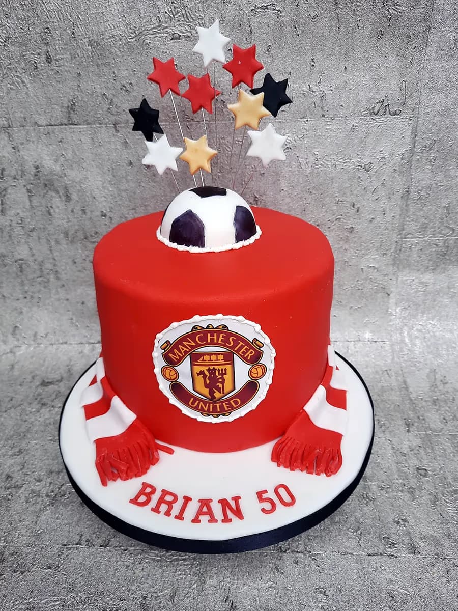 A round red birthday cake decorated with a Manchester United logo on the front, a half-sphere football on top, and an explosion of red, white, black, and gold stars on wires. A red and white striped scarf is draped around the base of the cake, and the words 'BRIAN 50' are written in red on the white cake board.