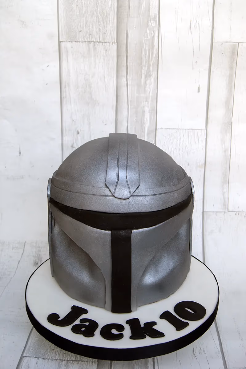A grey and black Mandalorian helmet cake on a white cake board, with 'Jack 10' written in black fondant.