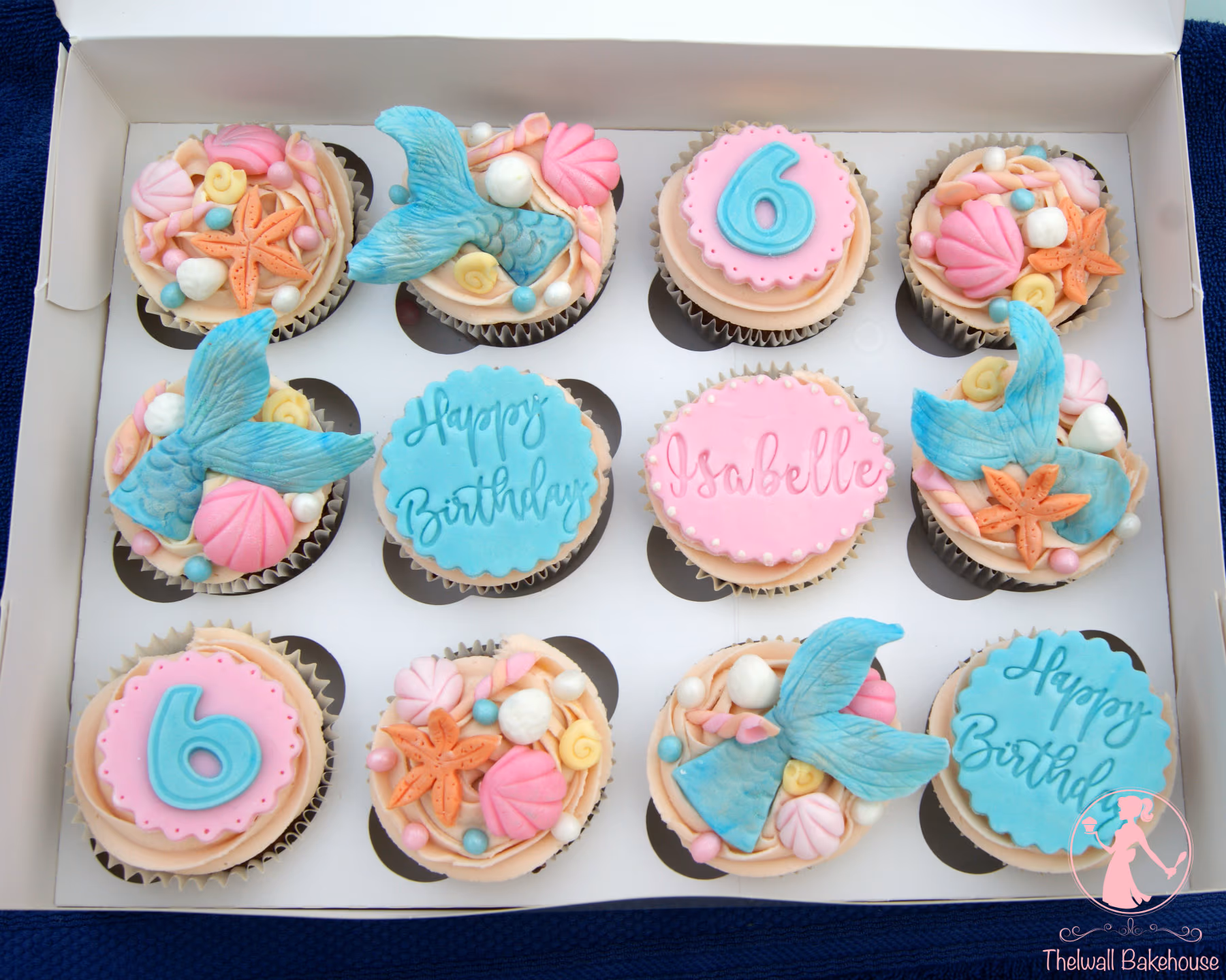 A dozen mermaid-themed cupcakes in a white box. The cupcakes have light peach frosting and are decorated with various fondant toppers, including blue mermaid tails, orange starfish, pink and yellow shells, and edible pearls. Some cupcakes feature pink fondant discs with a blue number '6' or the name 'Isabelle' embossed, while others have blue fondant discs with 'Happy Birthday' embossed.