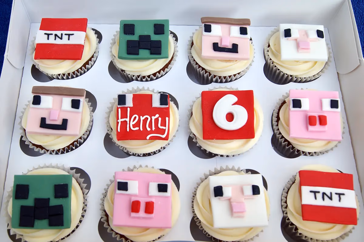 Twelve Minecraft-themed birthday cupcakes in a white box, featuring designs such as TNT blocks, Creeper faces, Steve faces, pigs, sheep, and personalized toppers for 'Henry' and the number '6'.