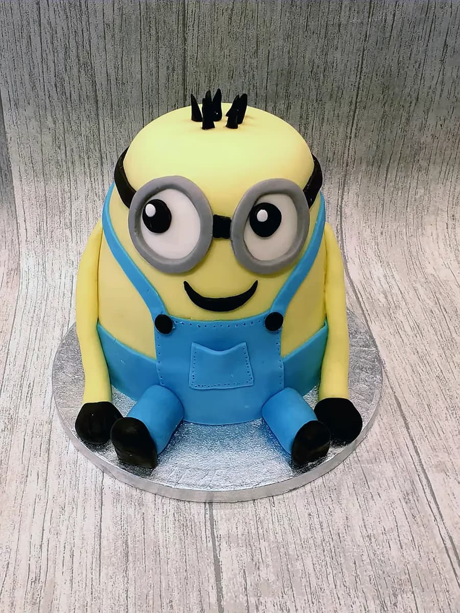 A Minion-shaped cake featuring a yellow body, blue overalls, goggles, and black hair, sitting on a silver cake board.