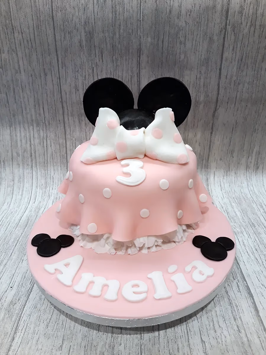 A single-tier pink Minnie Mouse themed birthday cake, decorated with white polka dots, a white bow with pink polka dots, and black Minnie Mouse ears on top. The number '3' is on the front, and the name 'Amelia' is written in white letters on the pink cake board, flanked by black Minnie Mouse head silhouettes.