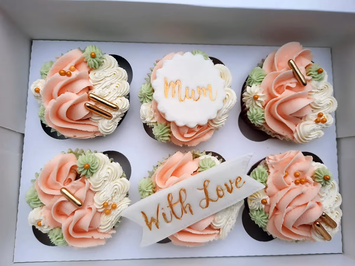 A white box containing six celebratory cupcakes, featuring pink, white, and light green piped frosting, adorned with gold sprinkles and edible gold bar decorations. One cupcake is topped with a white fondant disc bearing 'Mum' in gold, while another displays a white fondant banner with 'With Love' in gold.