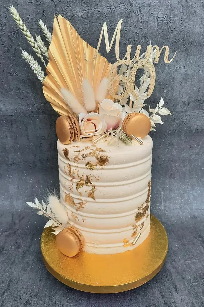 A single-tier cream-colored cake with horizontal ridges and gold leaf accents, topped with a gold 'Mum 60' topper, gold macarons, light pink roses, dried grasses, and a gold palm leaf fan, set on a gold cake board.