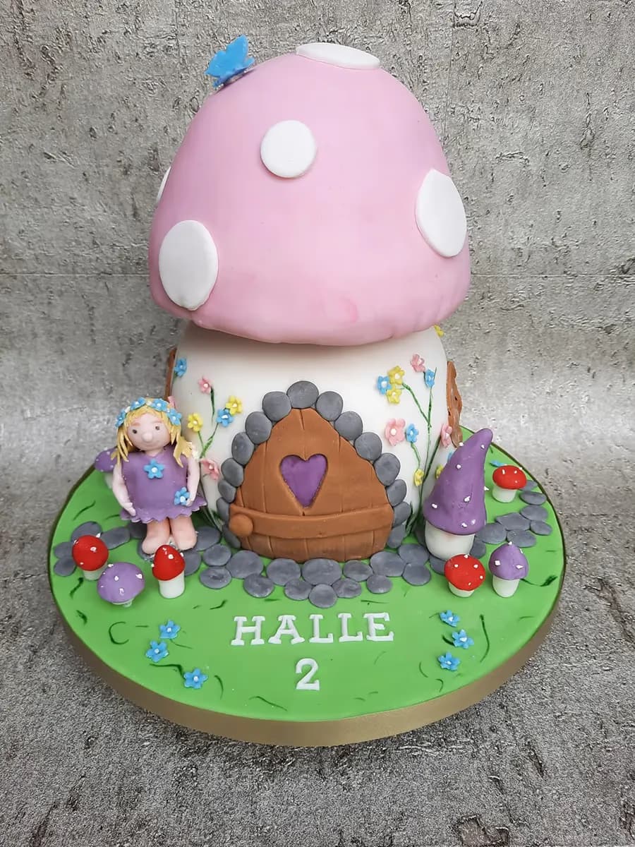 A whimsical two-tier mushroom house birthday cake with a pink cap and white base, decorated with colorful sugar flowers. A blonde fairy doll stands next to a wooden-style door with a purple heart. The green cake board features small red and purple toadstools and the name 'HALLE 2' in white lettering.