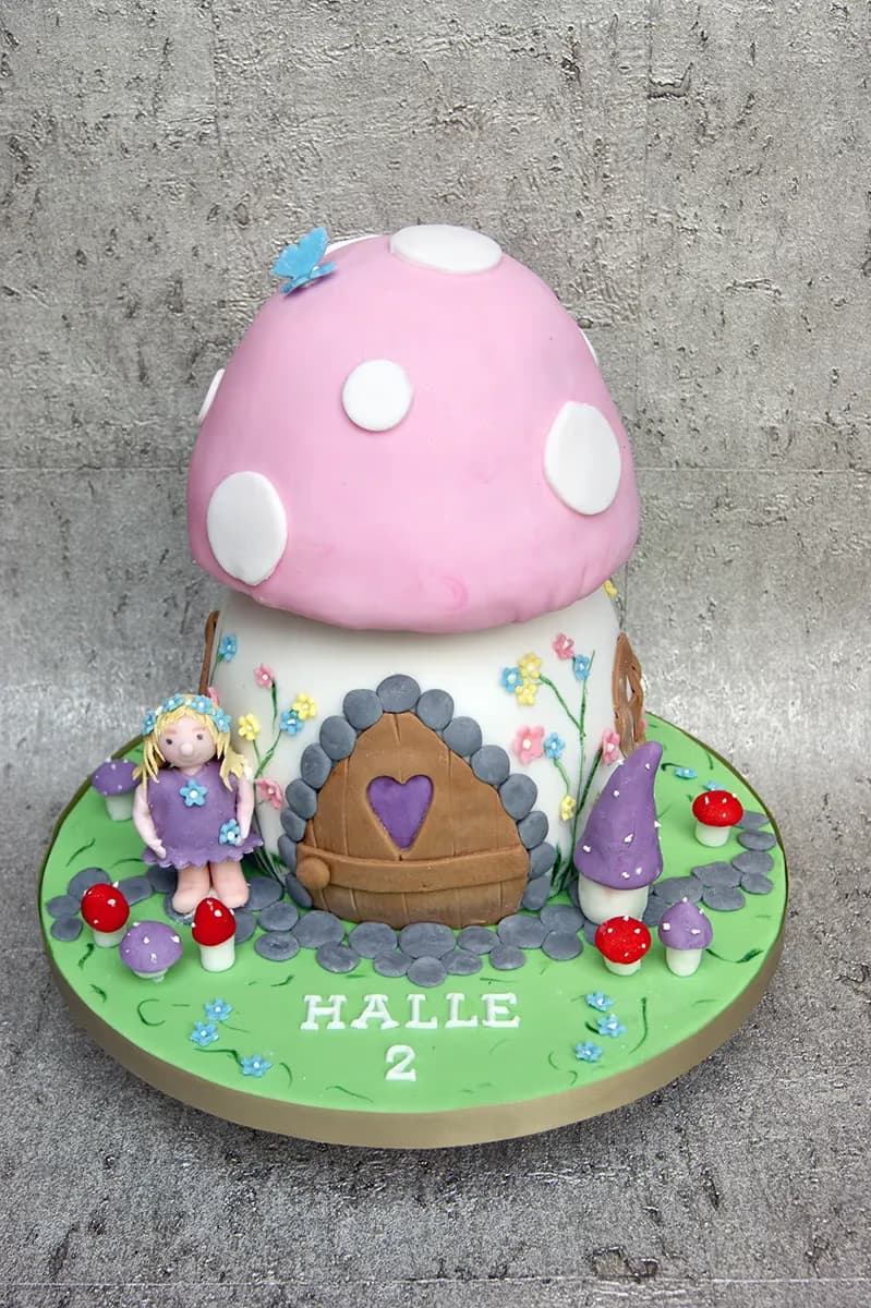 A whimsical mushroom house cake featuring a pink cap with white spots, a brown door with a heart, and a blonde fairy doll in a purple dress. The cake is surrounded by smaller red and purple mushrooms and tiny blue and pink flowers on a green base, with the name HALLE and the number 2 written on it.