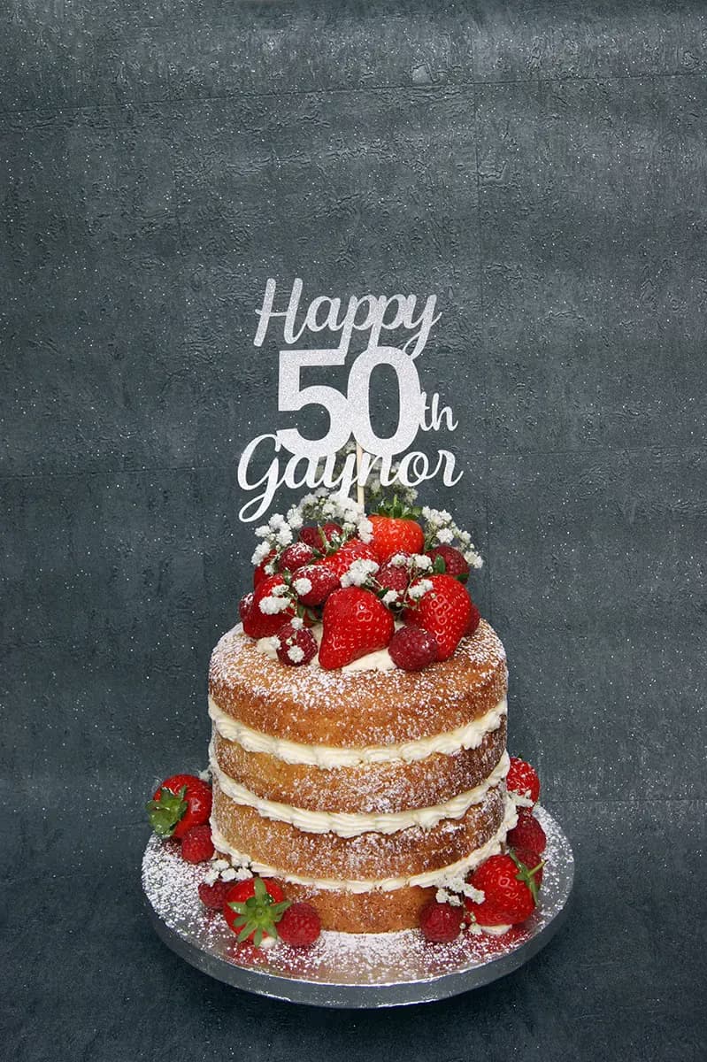 A three-tier naked birthday cake with white cream filling, topped with fresh strawberries, raspberries, and small white flowers, dusted with icing sugar, and featuring a silver glitter 'Happy 50th Gaynor' cake topper.