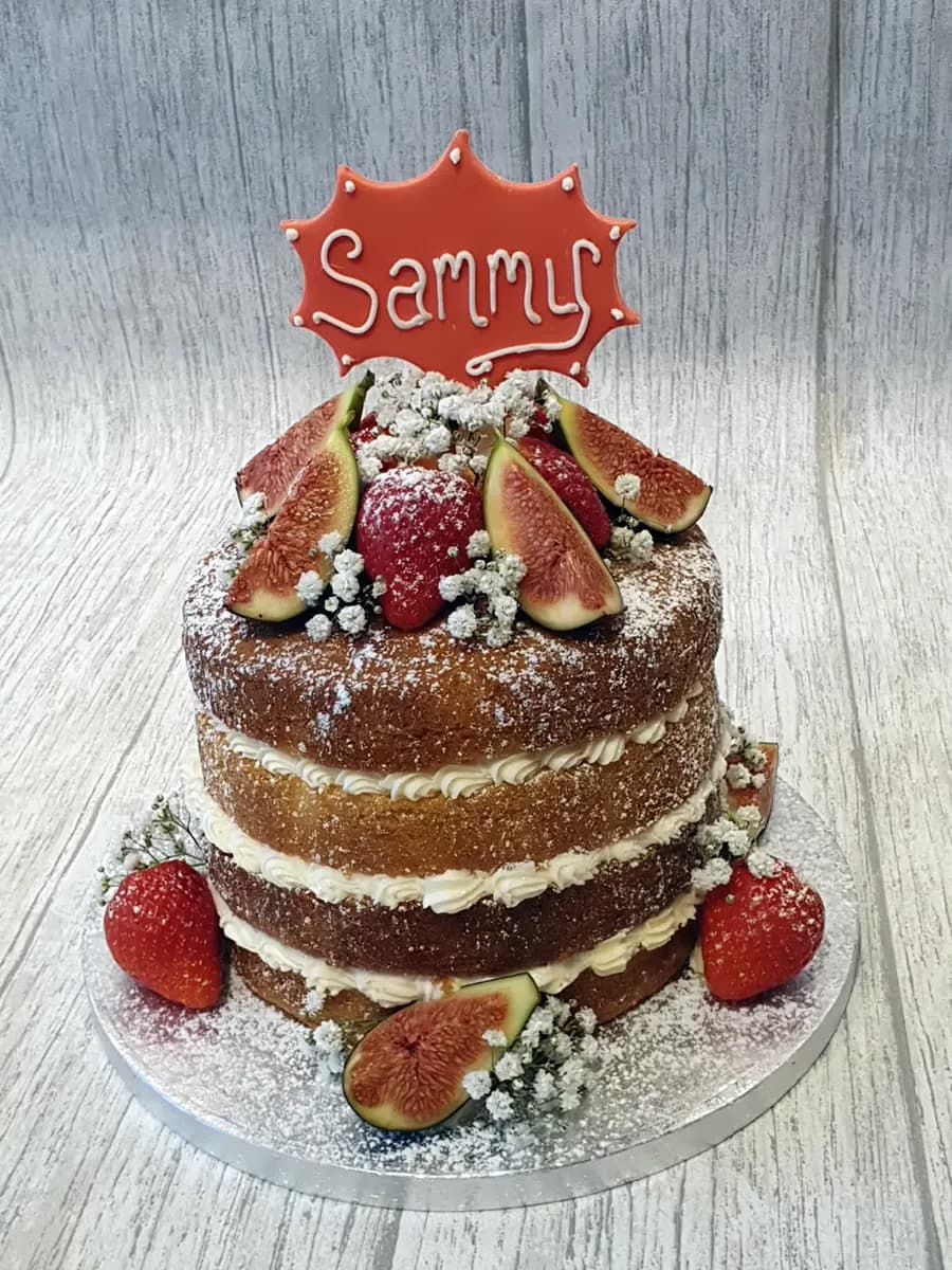 A three-tier naked cake with visible sponge layers and cream filling, adorned with fresh sliced figs, whole strawberries, white baby's breath flowers, a dusting of powdered sugar, and a red 'Sammy' name topper.