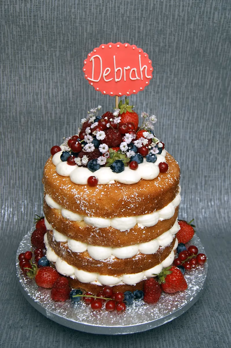 A three-tier naked sponge cake filled with white cream, dusted with powdered sugar, and decorated with an abundance of fresh mixed berries and small white flowers, topped with a red 'Debrah' cake topper.