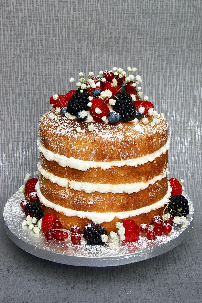 A three-tier naked cake decorated with fresh mixed berries, including raspberries, blackberries, blueberries, and redcurrants, along with small white flowers and a dusting of powdered sugar.
