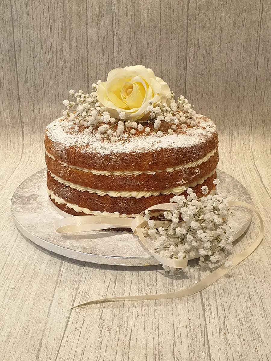 A two-tier naked cake dusted with icing sugar, decorated on top with a yellow rose and baby's breath flowers, and a cream ribbon tied around the base with additional baby's breath.