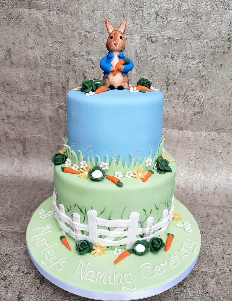 A two-tier Peter Rabbit themed naming ceremony cake, with a blue top tier and a light green bottom tier, decorated with carrots, cabbages, flowers, and a white picket fence, topped with a Peter Rabbit figurine holding a carrot. The cake board is inscribed with "Marley's Naming Ceremony".