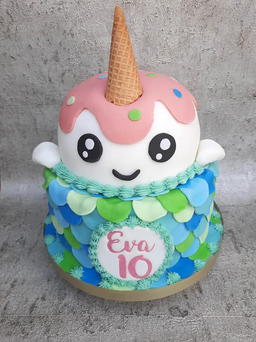 A two-tier birthday cake with a cute white narwhal character top tier, featuring a pink ice cream drip, colorful sprinkles, and a waffle cone horn, sitting on a bottom tier decorated with blue and green mermaid scales and a white plaque inscribed with 'Eva 10'.