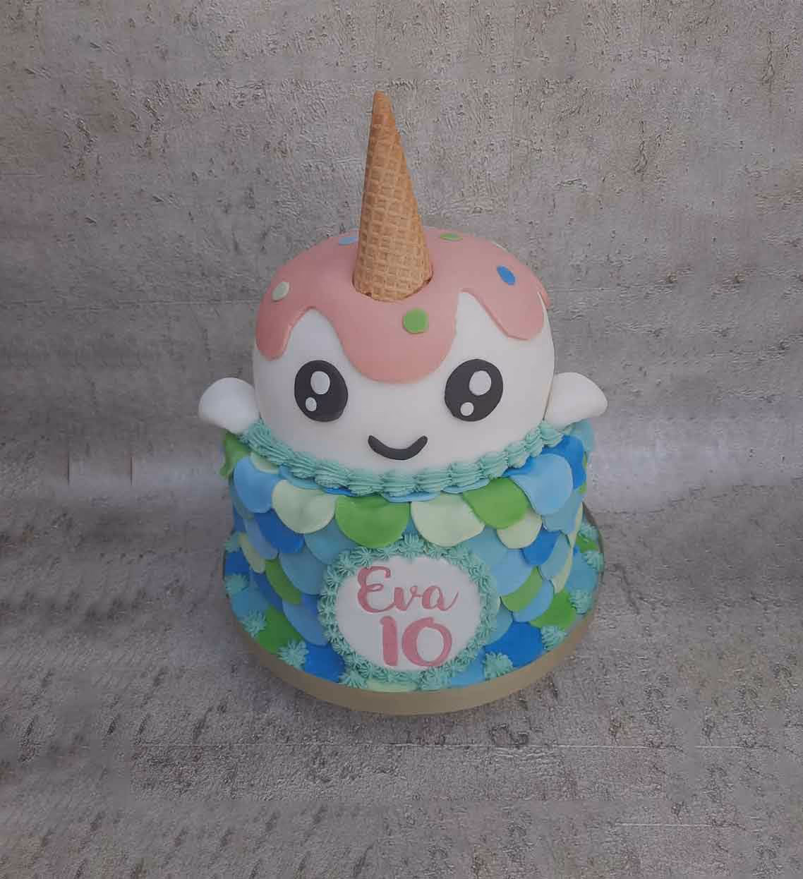 A whimsical two-tier birthday cake featuring a white, smiling narwhal or unicorn-like character with an ice cream cone horn and pink icing drip, sitting atop a bottom tier decorated with blue and green scales and a 'Eva 10' plaque.