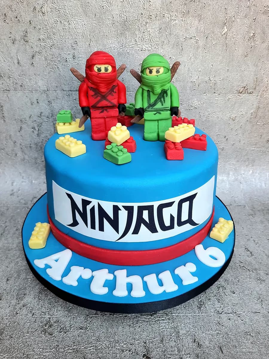 A vibrant blue, single-tier birthday cake decorated with red and green Lego Ninjago characters, surrounded by small edible red, yellow, and green Lego bricks. The side of the cake features a white band with 'NINJAGO' in black lettering, and the blue cake board has 'Arthur 6' written in white fondant letters.