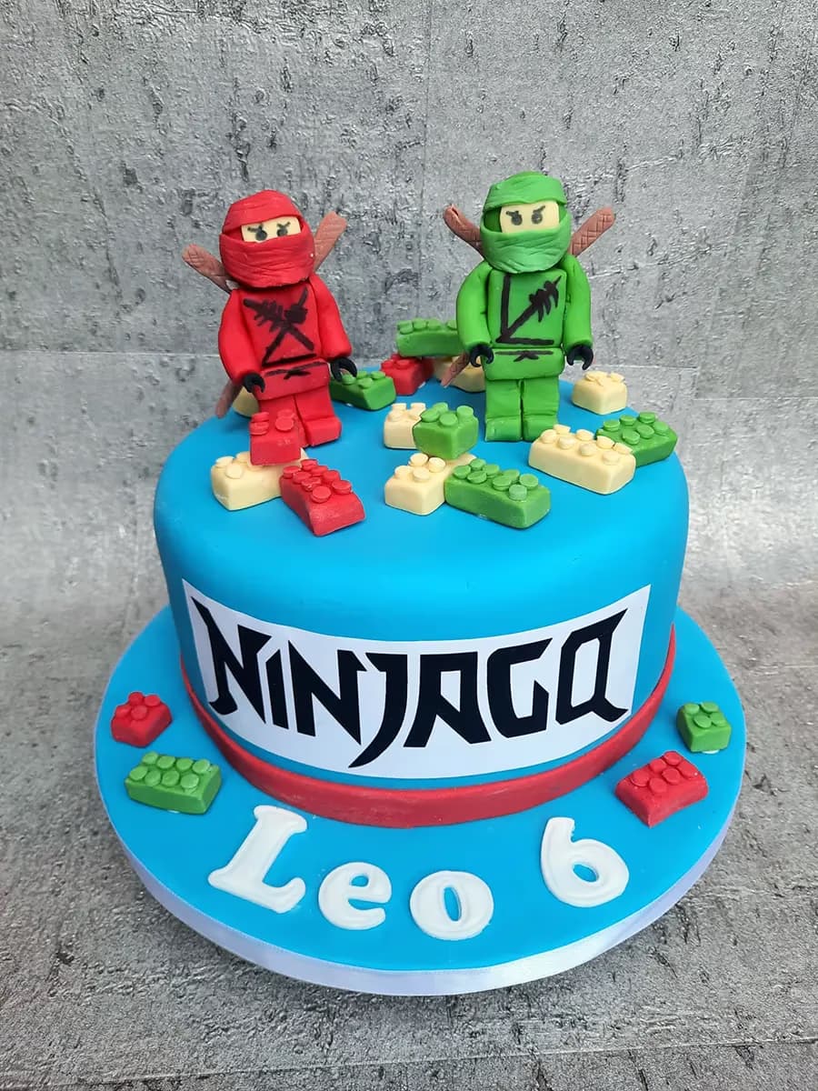 A round, bright blue fondant cake decorated with two fondant Lego Ninjago figures, one red and one green, standing on top. Around the figures are small fondant Lego bricks in red, green, and cream. The side of the cake features a white band with the word 'NINJAGO' in black letters, and on the cake board, in white letters, are the name 'Leo' and the number '6'.
